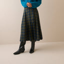 A person wearing a pleated plaid midi skirt in blue, green, and brown tones, paired with a bright teal sweater and black pointed-toe ankle boots, standing against a plain beige studio background.