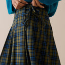 A close-up of a person wearing a pleated skirt with a green, blue, and black plaid pattern and a dark drawstring waist, paired with a teal sweater against a plain beige background.