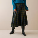 A person wearing a pleated plaid midi skirt in olive green, blue, and black, styled with a teal sweater and black knee-high boots against a neutral beige background.