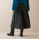 A person shown from the waist down wearing a pleated plaid skirt in olive green and blue, paired with a teal sweater and black boots, against a plain beige studio background.